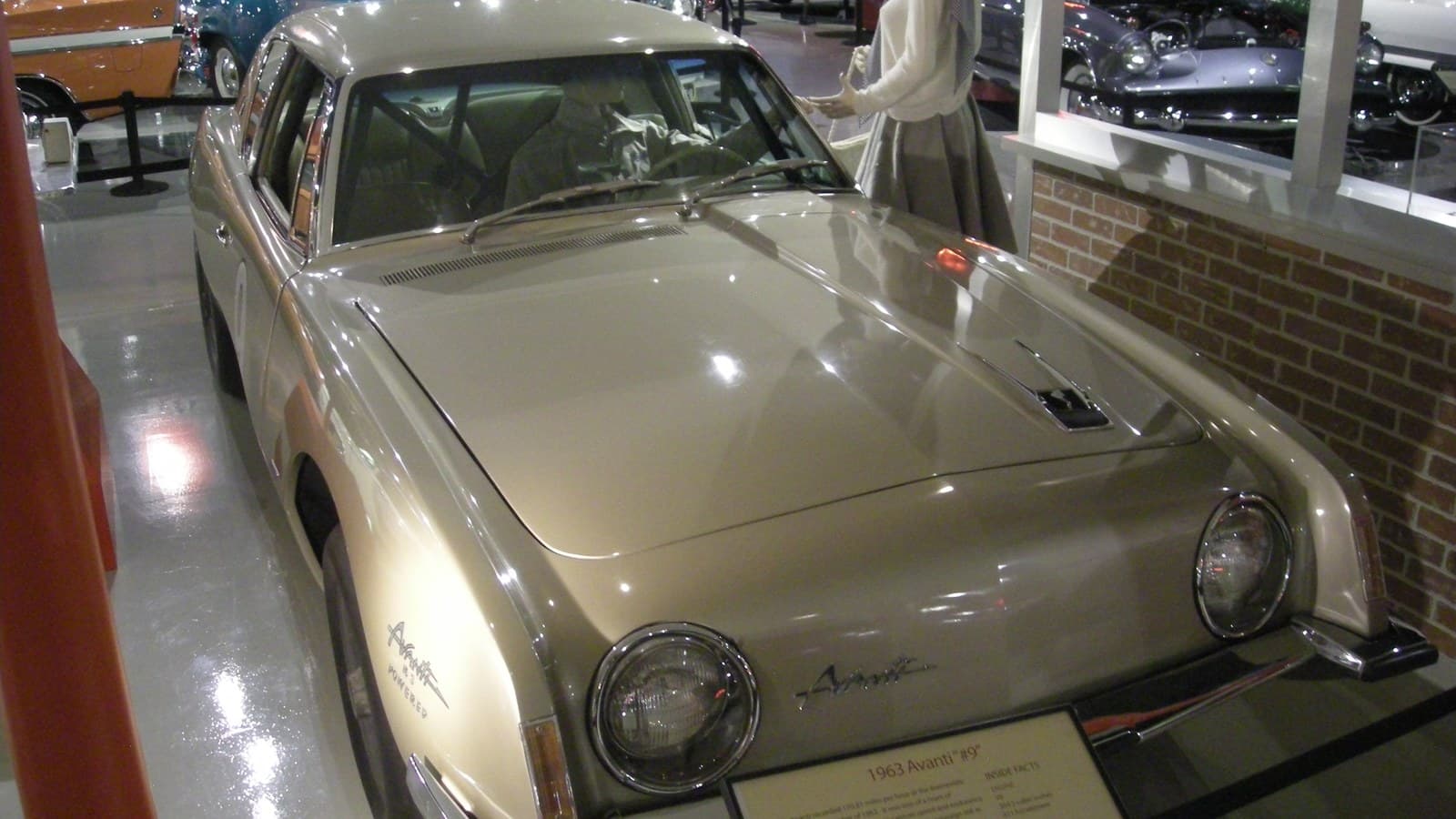 A gold 1963 Studebaker Avanti coupe parked indoors on a museum floor, seen from the front left, with round headlights, an Avanti script on the nose, R3 Powered lettering on the front fender, and a display placard reading 1963 Avanti #9 in front of the bumper.